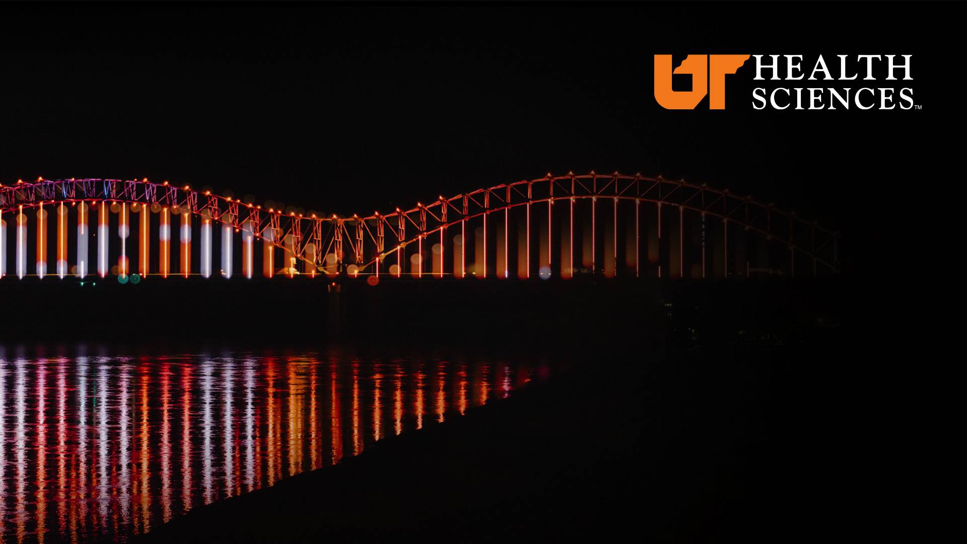 The Hernando De Soto Bridge at night and lite up with UT colors with UT Health Sciences logo.