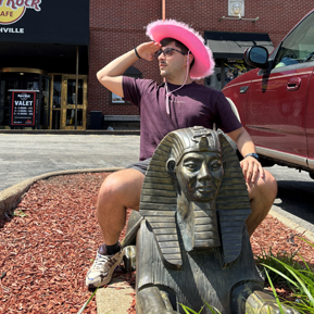 Male resident in a pink cowboy hat sitting on a sphynx statue in front of a Hard Rock Cafe