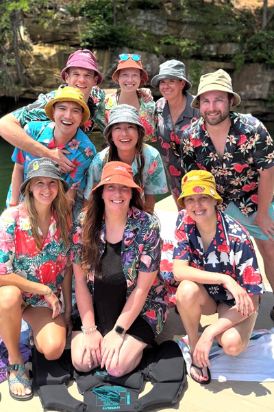 Eight fellows wearing colorful tropical shirts and bucket hats pose together outdoors near a rocky shoreline
