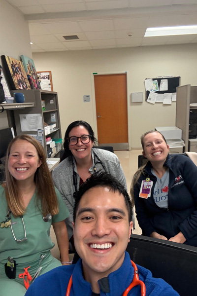 Four fellows gather for a selfie in a clinical workroom with desks, computers, and medical equipment