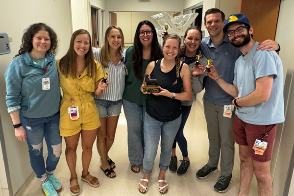 Fellows posing in a hallway with trophies and an award during a workplace celebration