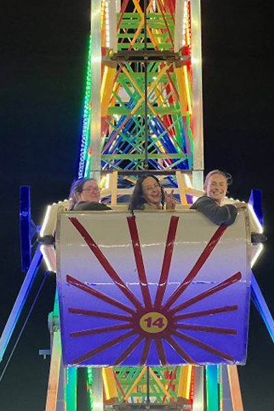 Three residents ride a brightly lit Ferris wheel gondola at a nighttime fair