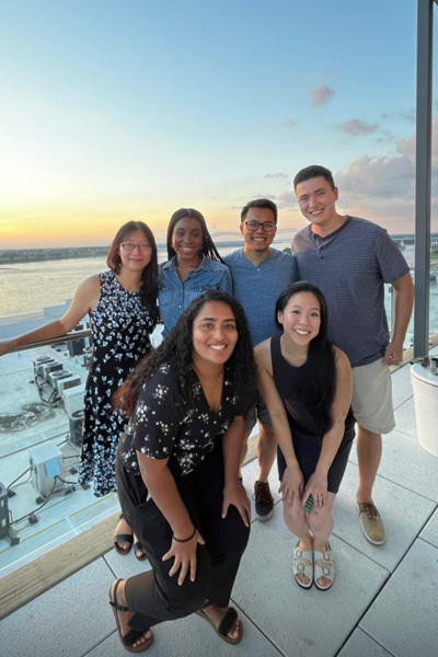 Six residents pose on a rooftop terrace overlooking a river and city skyline at sunset