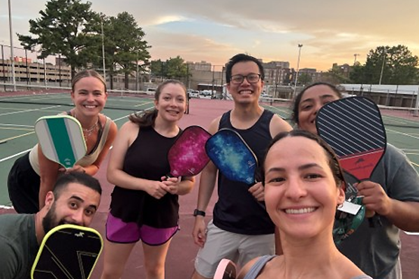Six residents standing on an outdoor pickleball court holding paddles at sunset
